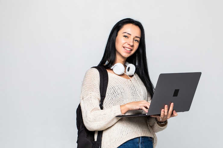 Foto gratuito mujer joven feliz que sostiene la computadora portátil y sobre la pared gris