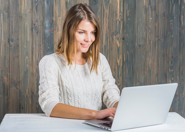 Mujer joven feliz que trabaja en la computadora portátil contra la pared de madera