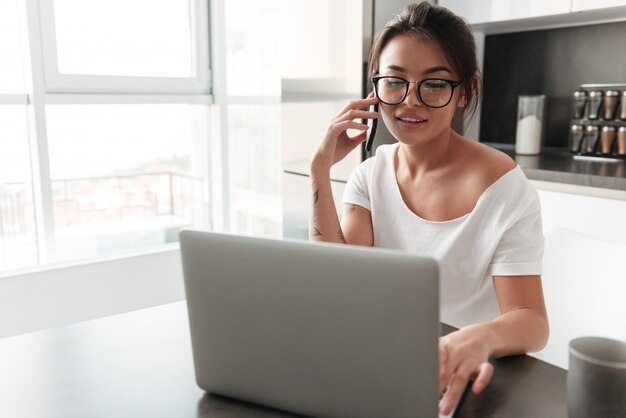 Mujer joven feliz que usa la computadora portátil que habla por el teléfono móvil.