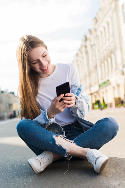 Mujer joven feliz que usa el smartphone que se sienta en la calle