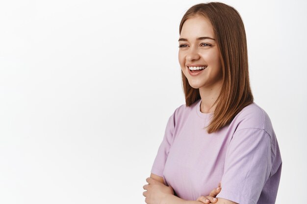 Mujer joven feliz riendo, sonriendo alegre, mirando hacia el lado izquierdo para su texto de promoción, anuncio, de pie en camiseta contra la pared blanca.