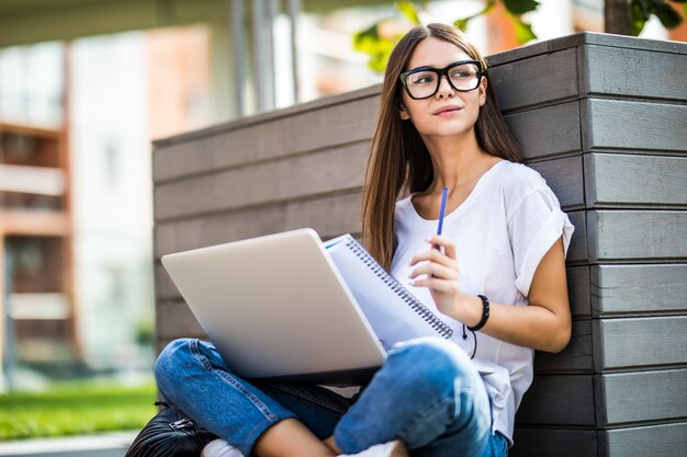 Mujer joven feliz en ropa casual y gafas usando una computadora portátil moderna y tomando notas mientras está sentado en el banco en la calle de la ciudad