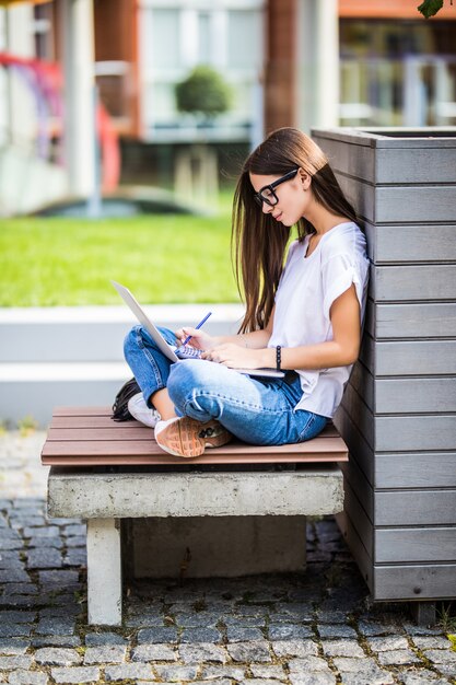 Mujer joven feliz en ropa casual y gafas usando una computadora portátil moderna y tomando notas mientras está sentado en el banco en la calle de la ciudad