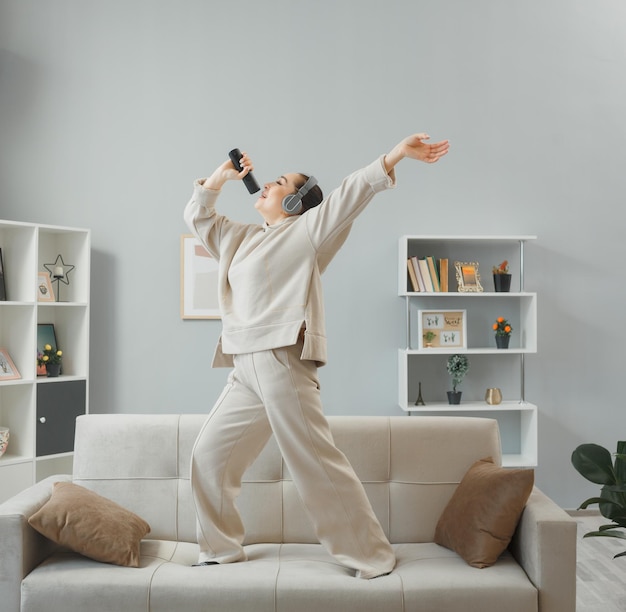 Mujer joven feliz con ropa informal con auriculares bailando en un sofá en el interior de la casa sosteniendo el mando a distancia usándolo como micrófono cantando divirtiéndose