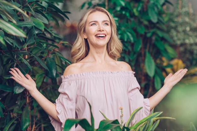 Mujer joven feliz rubia despreocupada que se coloca cerca de las plantas que se encogen sus manos