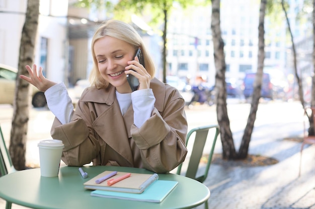 Foto gratuita mujer joven feliz saliente sentada en una cafetería al aire libre hablando por teléfono móvil sonriendo pasando el tiempo