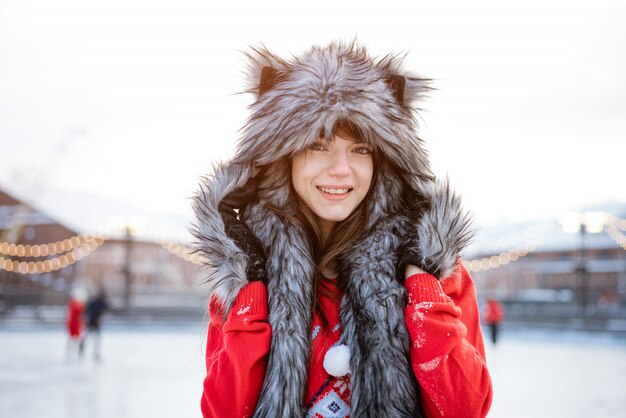 Mujer joven feliz con un sombrero de lobo en invierno en la pista de hielo posa con un suéter rojo afuera por la tarde