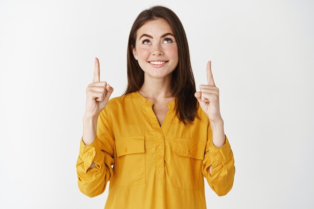 Mujer joven feliz sonriendo, señalando y mirando la oferta promocional, el concepto de compras y publicidad, de pie sobre una pared blanca.