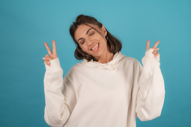 Mujer joven feliz en sudadera con capucha blanca posando alegremente en azul en el estudio