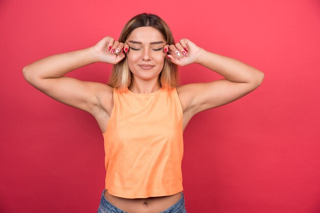 Mujer joven feliz tocando sus ojos en la pared roja.