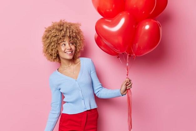 La mujer joven feliz usa un suéter casual y los pantalones sostienen un montón de globos inflados en forma de corazón que están de buen humor tiene un estado de ánimo festivo aislado sobre fondo rosa Concepto de personas y vacaciones