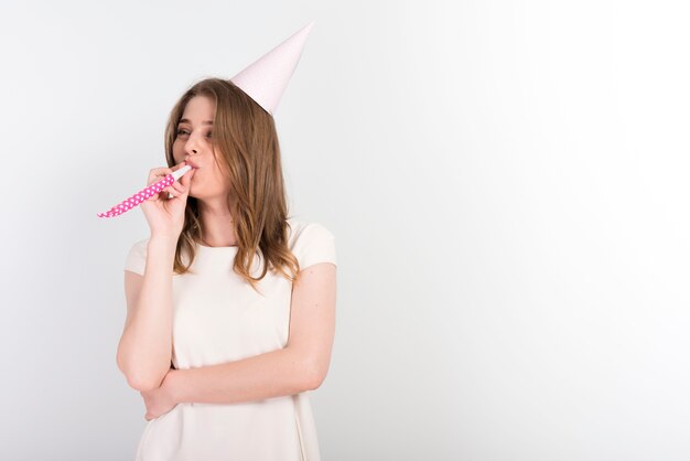 Mujer joven festiva en el sombrero de fiesta soplando ruido