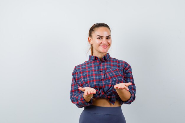 Mujer joven fingiendo sostener algo en camisa a cuadros, pantalones y mirando feliz, vista frontal.