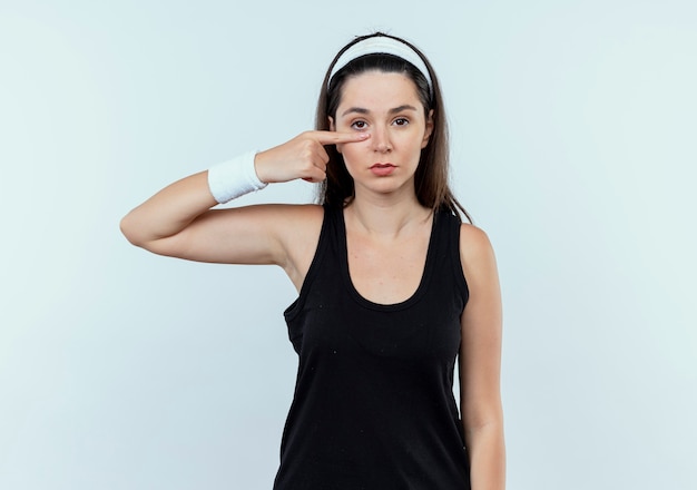 Mujer joven fitness en diadema apuntando con el dedo a la nariz con rostro serio mirando a la cámara de pie sobre fondo blanco.