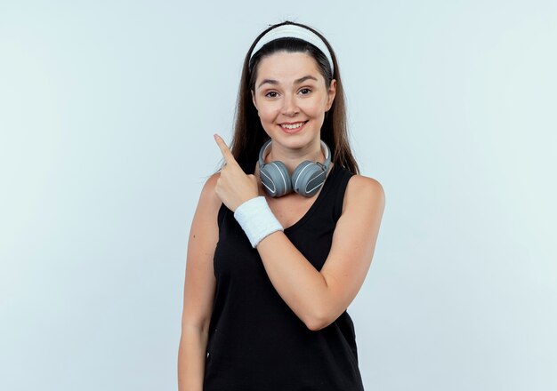 Mujer joven fitness en diadema con auriculares apuntando con el dedo hacia el lado sonriendo con cara feliz de pie sobre la pared blanca