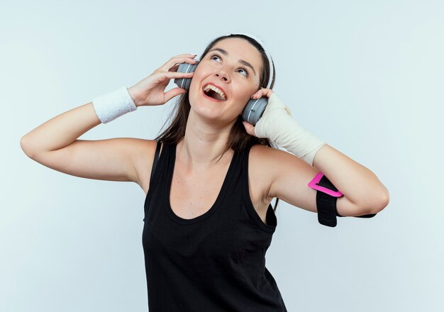 Mujer joven fitness en diadema con auriculares y brazalete de smartphone disfrutando de su música favorita de pie sobre fondo blanco.