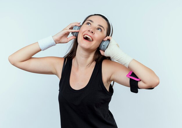 Mujer joven fitness en diadema con auriculares y brazalete de smartphone disfrutando de su música favorita de pie sobre la pared blanca