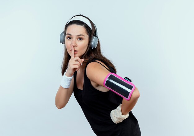 Mujer joven fitness en diadema con auriculares y brazalete de smartphone haciendo gesto de silencio con el dedo en los labios de pie sobre fondo blanco.