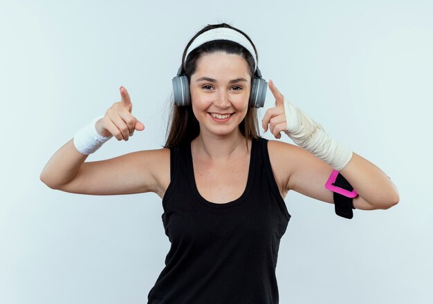 Mujer joven fitness en diadema con auriculares y brazalete de teléfono inteligente mirando a la cámara sonriendo con cara feliz apuntando con los dedos índices al lado de pie sobre fondo blanco