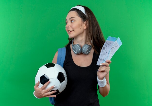 Mujer joven fitness en diadema con auriculares y mochila sosteniendo un balón de fútbol y boletos de avión mirando a un lado sonriendo de pie sobre la pared verde