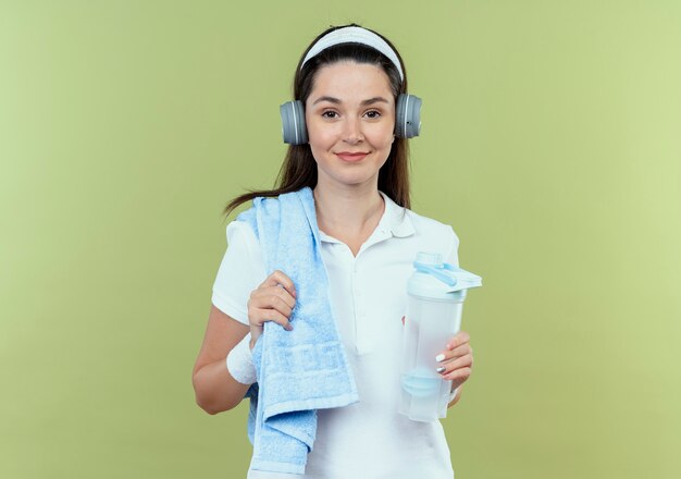 Mujer joven fitness en diadema con auriculares y toalla en el hombro sosteniendo una botella de agua mirando a la cámara sonriendo de pie sobre fondo claro