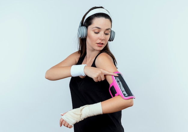 Mujer joven fitness en diadema con auriculares tocando el brazalete de su smartphone mirando confiado de pie sobre fondo blanco.