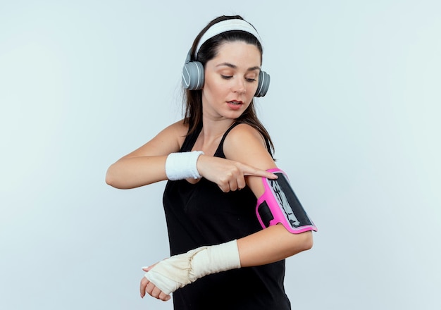 Foto gratuita mujer joven fitness en diadema con auriculares tocando el brazalete de su smartphone mirando confiado de pie sobre la pared blanca