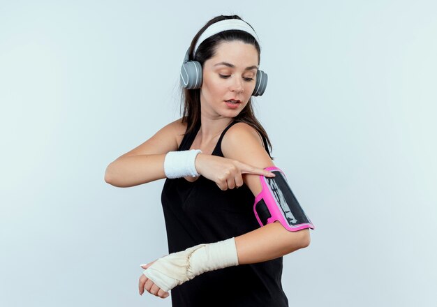 Mujer joven fitness en diadema con auriculares tocando el brazalete de su smartphone mirando confiado de pie sobre la pared blanca