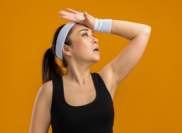 Mujer joven fitness con diadema y brazaletes mirando hacia arriba con la mano en la cabeza está cansado y con exceso de trabajo de pie sobre la pared naranja