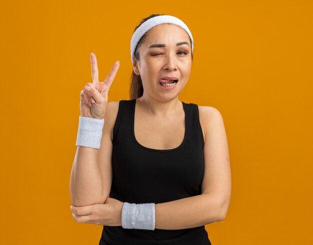 Mujer joven fitness con diadema y brazaletes sonriendo y guiñando un ojo mostrando v-sign de pie sobre la pared naranja