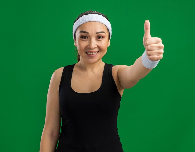 Mujer joven fitness con diadema y brazaletes con una sonrisa en la cara mostrando los pulgares para arriba de pie sobre la pared verde