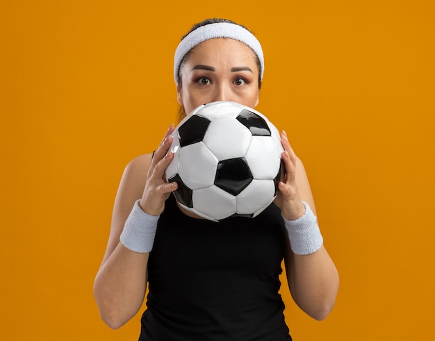 Foto gratuita mujer joven fitness con diadema y brazaletes sosteniendo un balón de fútbol delante de su rostro preocupado de pie sobre la pared naranja