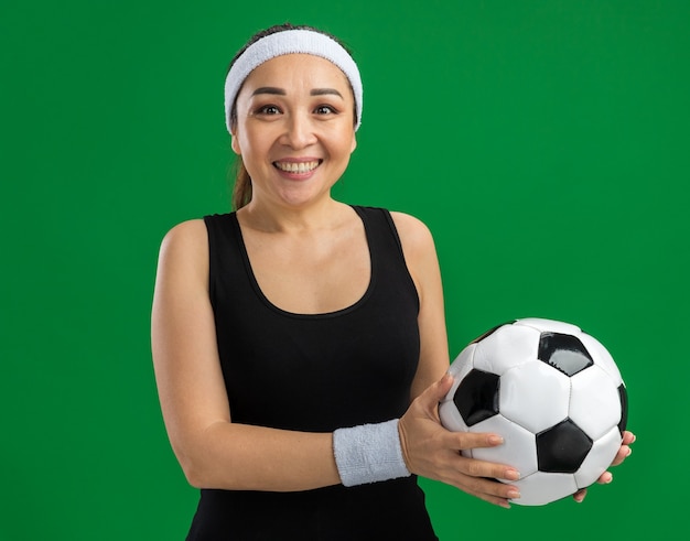 Mujer joven fitness con diadema y brazaletes sosteniendo un balón de fútbol sonriendo feliz y positivo