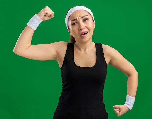Mujer joven fitness con diadema y brazaletes tensos y seguros levantando puños mostrando fuerza y poder de pie sobre la pared verde