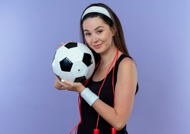 Mujer joven fitness en diadema con cuerda alrededor del cuello sosteniendo un balón de fútbol mirando a la cámara sonriendo de pie sobre fondo azul.