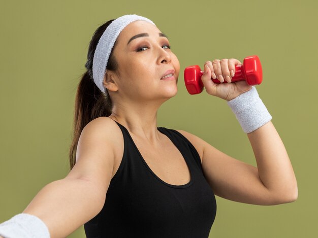 Mujer joven fitness con diadema con mancuernas haciendo ejercicios con expresión de confianza de pie sobre la pared verde