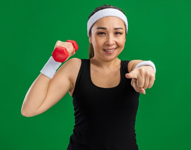 Mujer joven fitness con diadema con mancuernas haciendo ejercicios sonriendo confiado apuntando con el dedo índice de pie sobre la pared verde