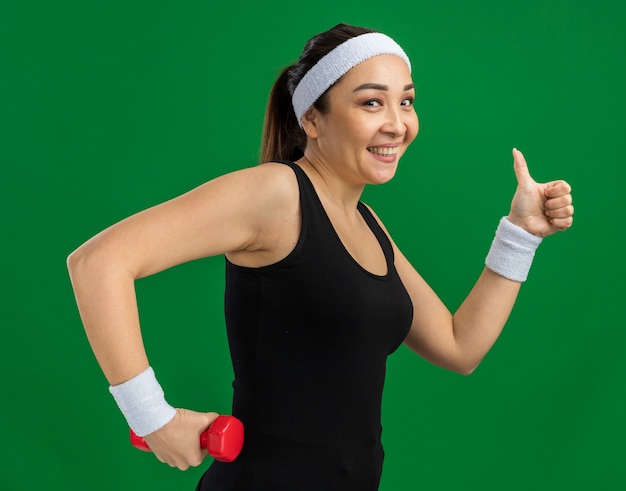 Mujer joven fitness con diadema con mancuernas haciendo ejercicios sonriendo mostrando los pulgares para arriba de pie sobre la pared verde