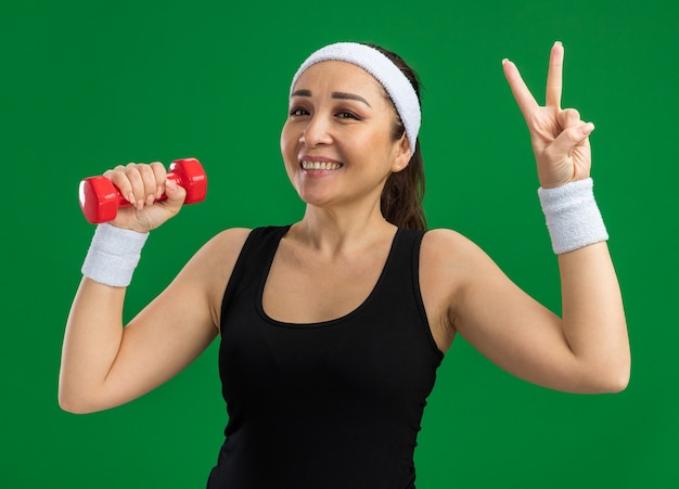 Mujer joven fitness con diadema con mancuernas haciendo ejercicios sonriendo mostrando v-sign de pie sobre la pared verde