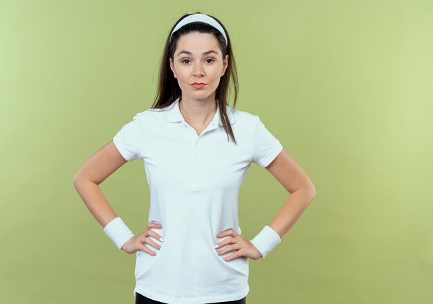 Mujer joven fitness en diadema mirando a la cámara con expresión de confianza con los brazos en la cadera de pie sobre fondo claro