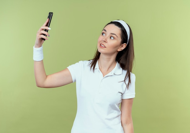 Mujer joven fitness en diadema mirando confundido tomando selfie con smartphone parado sobre pared ligera