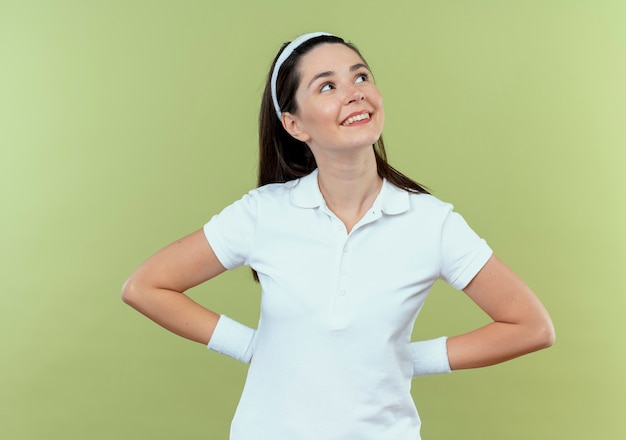 Foto gratuita mujer joven fitness en diadema mirando a un lado con expresión de confianza sonriendo de pie sobre fondo claro