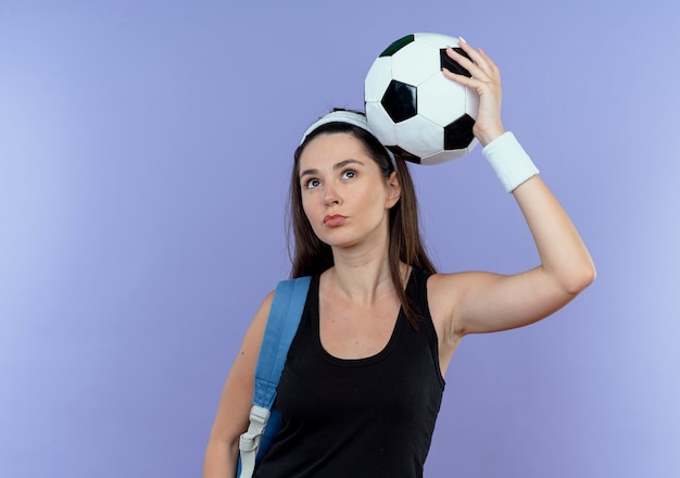 Mujer joven fitness en diadema con mochila sosteniendo un balón de fútbol sobre la cabeza mirando confiado de pie sobre fondo azul.