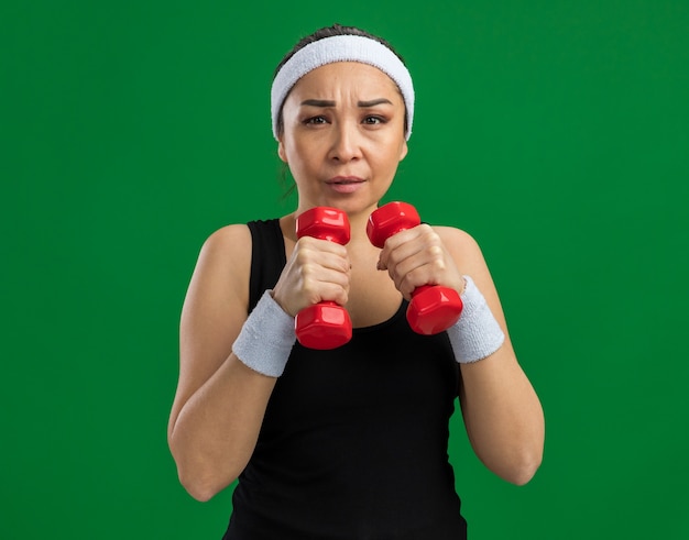 Mujer joven fitness con diadema con pesas haciendo ejercicios tensos y seguros de pie sobre la pared verde