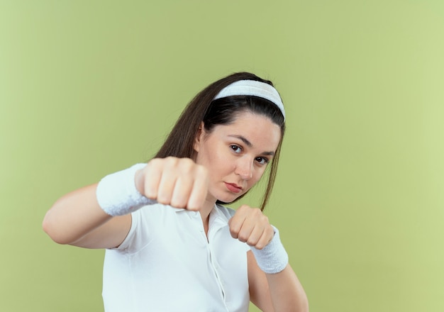 Mujer joven fitness en diadema posando como un boxeador apuntando con el puño a la cámara de pie sobre fondo claro