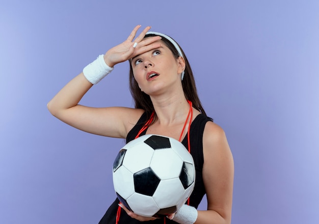 Mujer joven fitness en diadema con saltar la cuerda alrededor del cuello sosteniendo un balón de fútbol mirando hacia arriba cansado de pie sobre fondo azul.