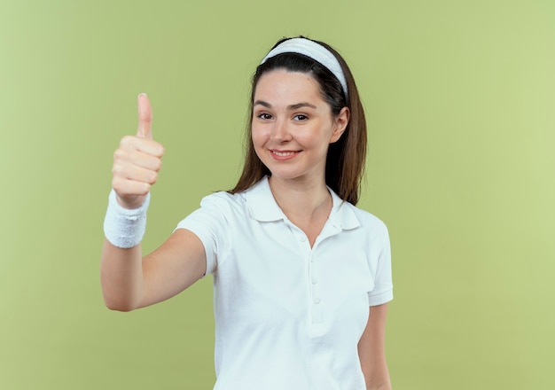 Mujer joven fitness en diadema sonriendo alegremente mostrando los pulgares para arriba de pie sobre la pared de luz