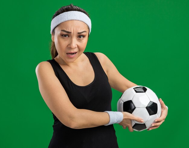 Mujer joven fitness con diadema sosteniendo un balón de fútbol con cara seria