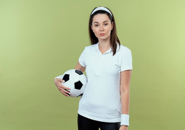 Mujer joven fitness en diadema sosteniendo un balón de fútbol mirando a la cámara con seria expresión de confianza de pie sobre fondo claro