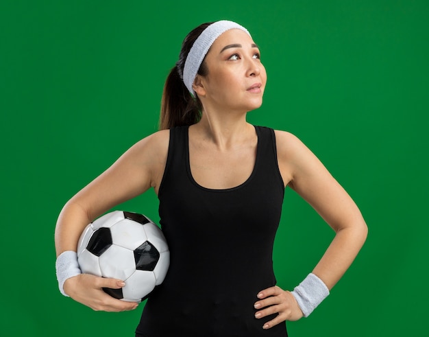 Mujer joven fitness con diadema sosteniendo un balón de fútbol mirando a un lado con una sonrisa en la cara de pie sobre la pared verde